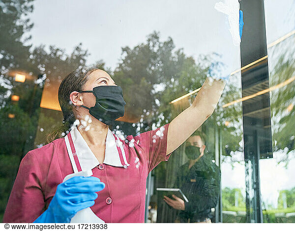 Female hotel maid in face mask cleaning hotel window