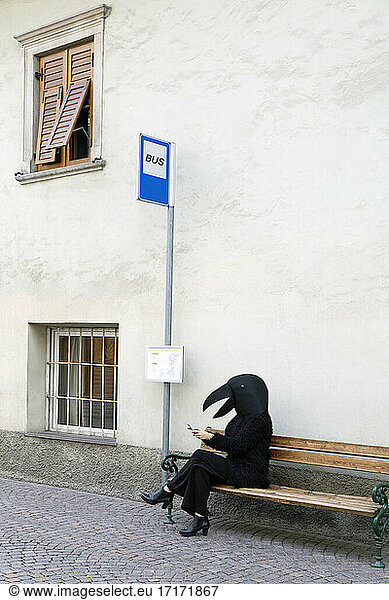 Female in crow costume using mobile phone while sitting on bench of bus stop