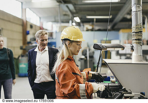 Female industrial worker in hardhat working on machinery while managers in background at factory