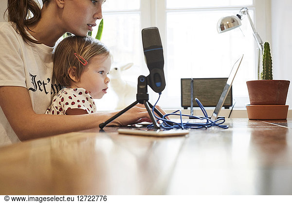 Female influencer sitting with daughter while marketing through laptop at home