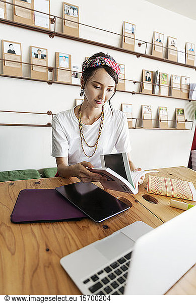 Female Japanese professional sitting at table in a co-working space  using digital tablet and laptop  holding book.