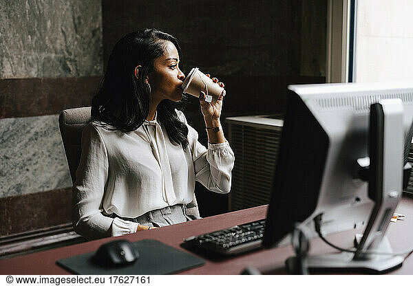 Female lawyer drinking coffee from disposable cup while sitting at desk in office