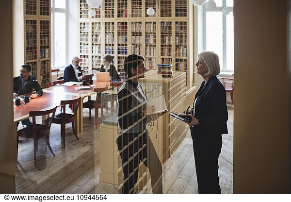 Female lawyers discussing while standing seen through glass in library