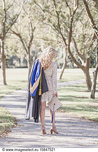Female long blond curly hair holds graduation cap and gown on path