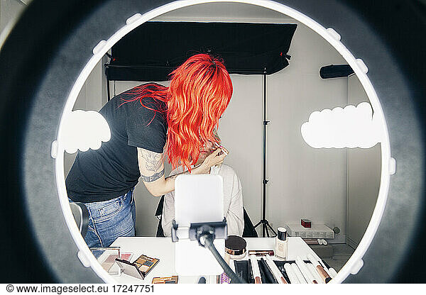 Female make-up artist applying make-up to model seen through ring light at studio