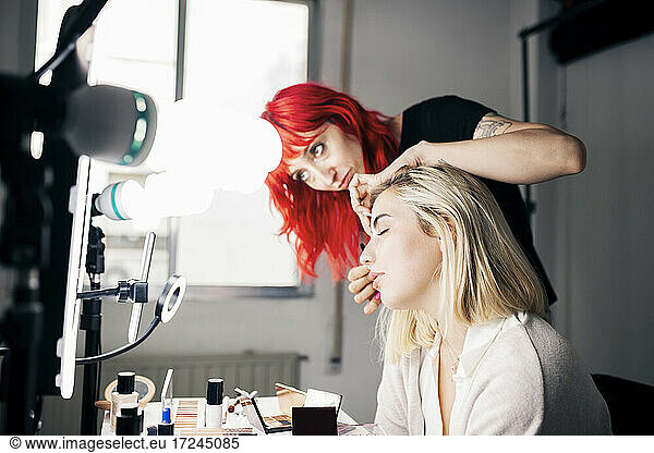 Female make-up artist applying mascara to model during vlogging through smart phone at studio