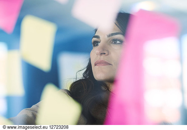 Female mature engineer reading adhesive notes seen through glass in office