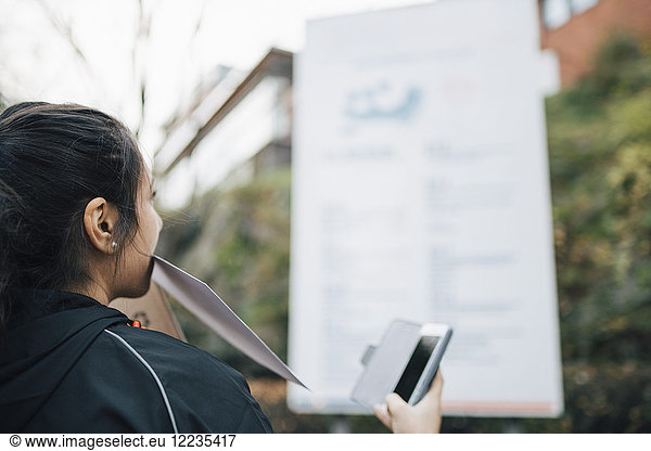 Female messenger carrying document in mouth and holding smart phone looking at billboard