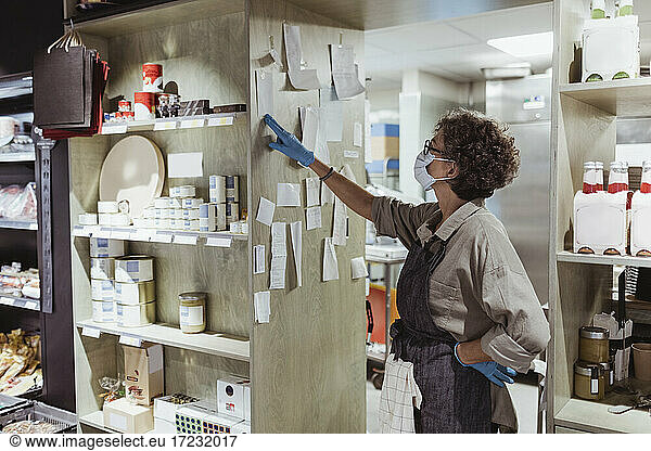 Female owner looking at adhesive notes stick on rack at delicatessen shop during pandemic