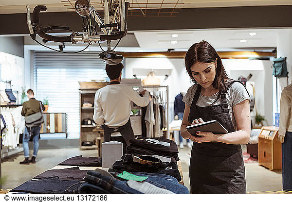 Female owner using digital tablet while coworker arranging clothes on rack at store