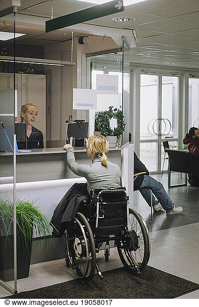 Female patient sitting on wheelchair and doing inquiry with receptionist at healthcare center