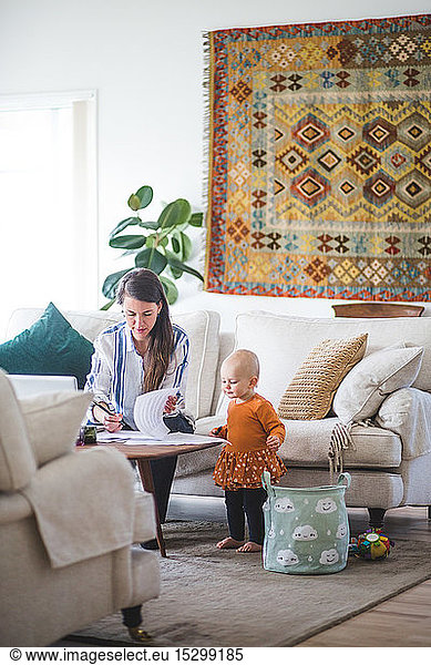 Female professional freelancing while daughter looking at papers on table in living room