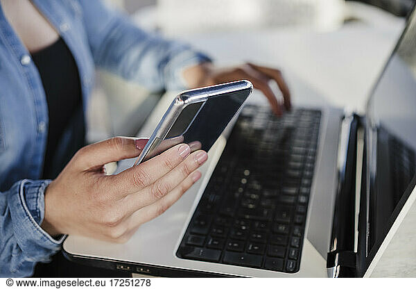 Female professional holding mobile phone while working on laptop in cafe