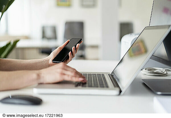 Female professional holding mobile phone while working on laptop in office