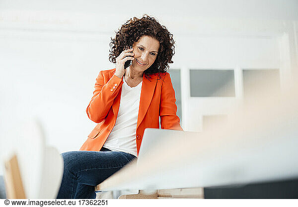 Female professional using laptop while talking on mobile phone at office