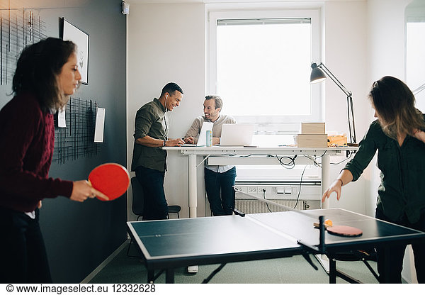 Female professionals playing table tennis while male colleagues using laptop at creative office
