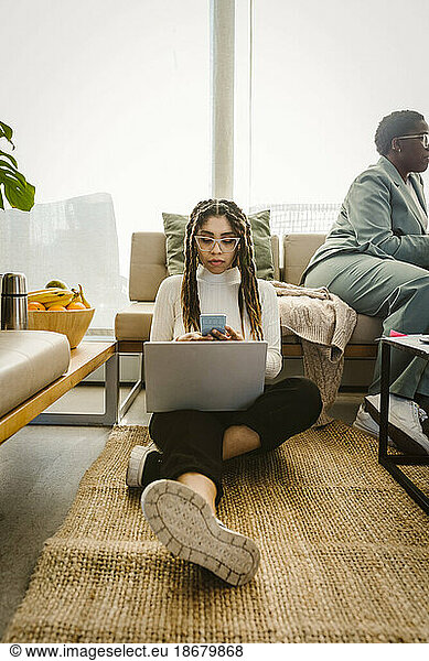 Female programmer using smart phone while sitting with laptop on carpet in office