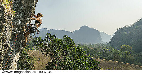 Female rock climber high on wall with trees and mountains behind  Asia