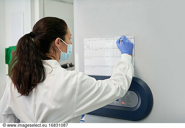 Female scientist in face mask marking schedule on wall in laboratory