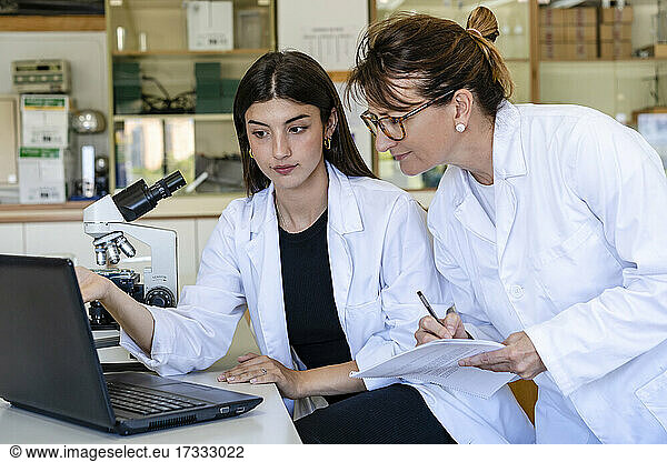 Female scientist writing on document while discussing with colleague over laptop