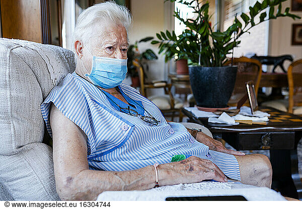 Female senior with protective mask sitting in armchair at home
