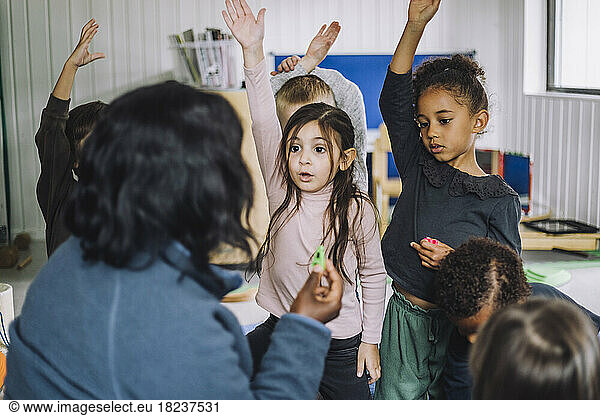 Female teacher asking questions and students raising hand to answer at day care center