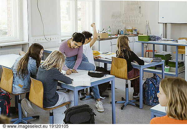 Female teacher assisting schoolgirls while sitting on desk in classroom