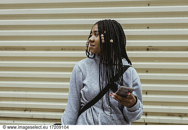 Female teenager with smart phone looking away against metal wall