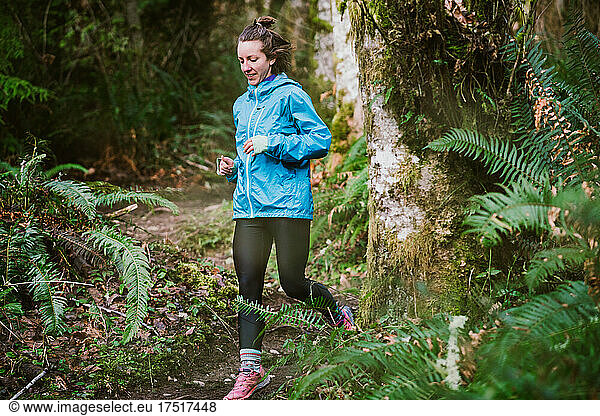 Female trail runner in blue jacket runs in lush fern forest