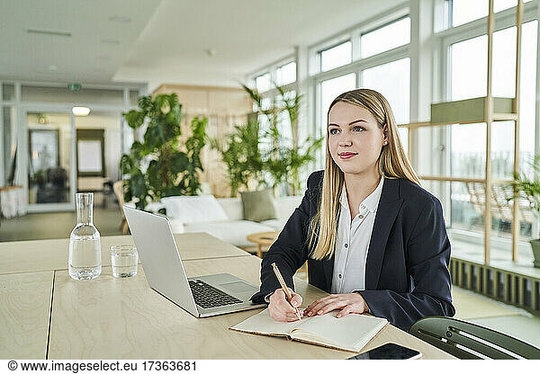 Female trainee looking away while sitting with diary and laptop at desk in office