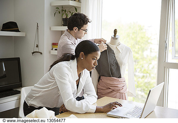 Female working on laptop while colleague pinning dress on model in studio
