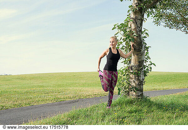 Fit female athlete stretching leg near tree during training in meadow