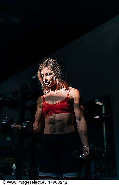 fit woman using dumbbells in dark gym with sun on her face