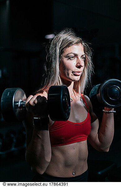 fit woman using dumbbells in dark gym with sun on her face