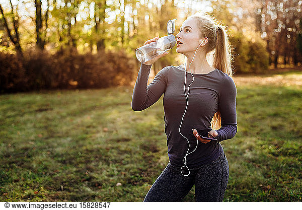 Fitness in the park  girl drinking water  holding smartphone and headphones in her hand.