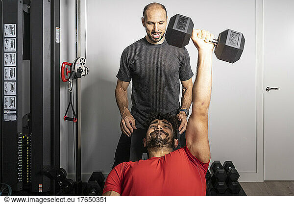 Fitness instructor motivating young man lifting dumbbell at gym