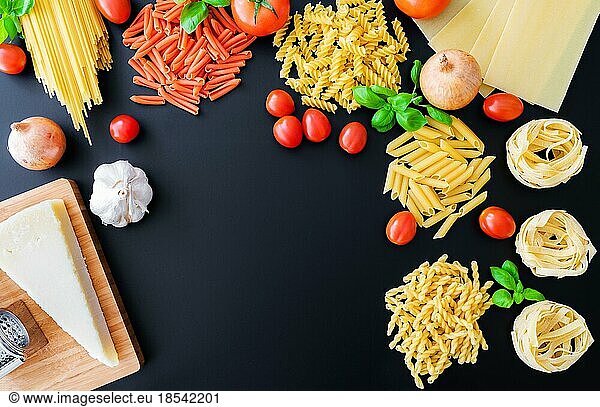 flat lay of different uncooked italian pasta noodles on dark background with basil leaves  fresh tomatoes and onions  parmesan cheese and grater