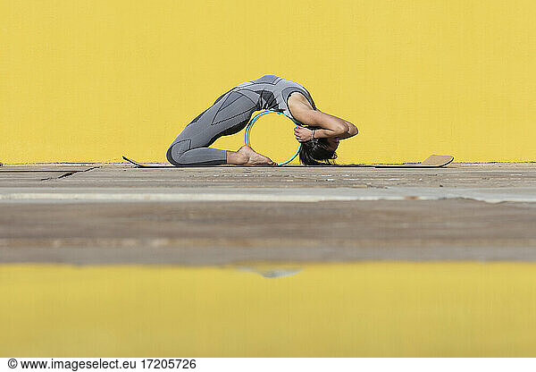 Flexible woman with yoga wheel bending over backwards by yellow wall
