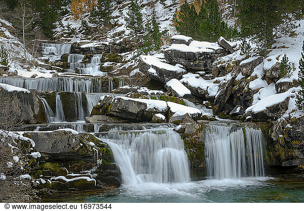 Fluss  der durch ein verschneites Tal mit Wasserfällen und verschneiten Bäumen fließt