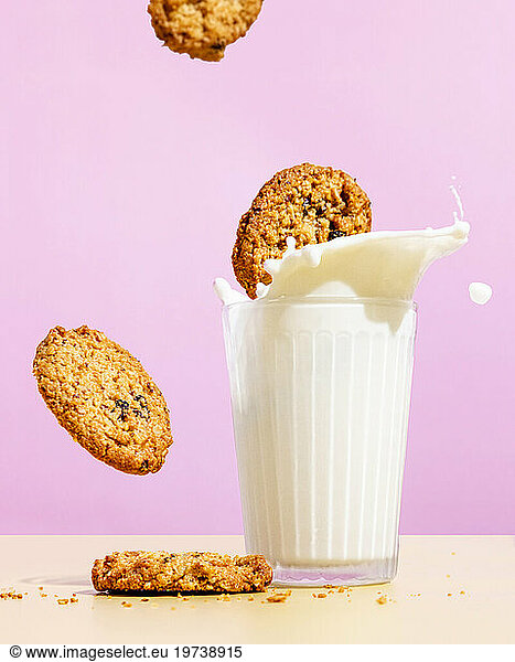 Flying oatmeal cookies near milk splashing in glass against pink colored background