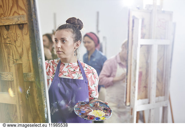 Focused female artist with palette painting at easel in art class studio
