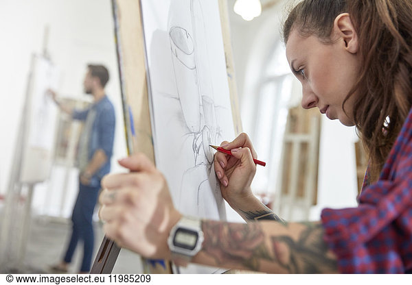 Focused female artist with tattoo sketching at easel in art class studio