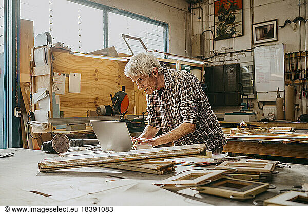 Focused senior male carpenter working on laptop at repair shop