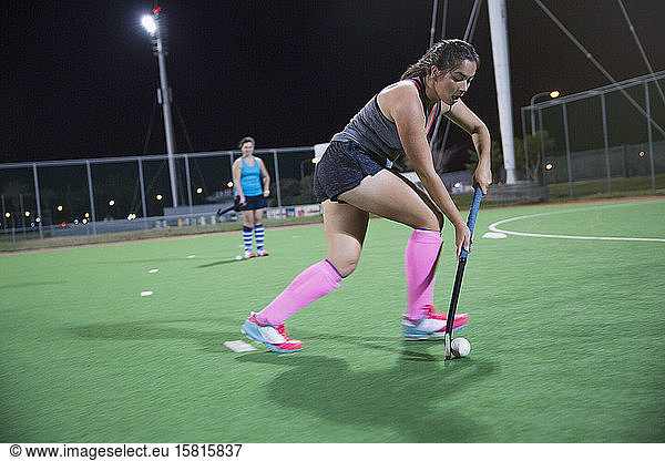 Focused young female field hockey player playing on field at night