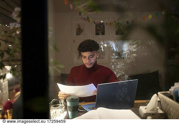 Focused young man with paperwork working from home at laptop at night