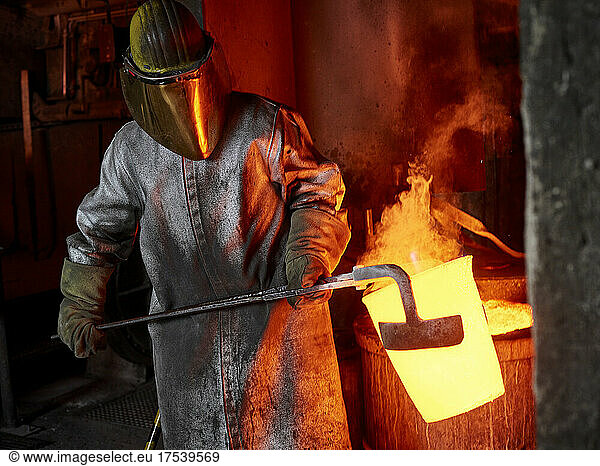 Foundry worker carrying burning container with hand tool in industry