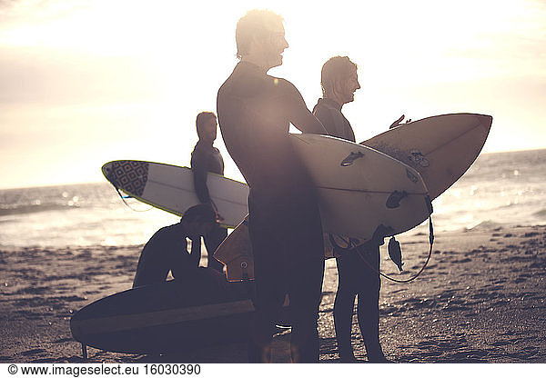Four men wearing wetsuits standing on a sandy beach  carrying surfboards.
