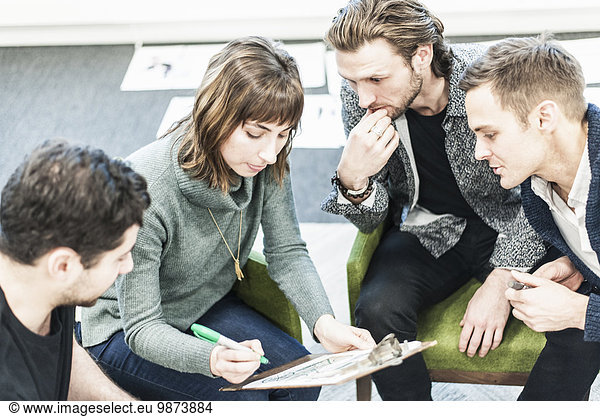 Four people  colleagues at a meeting  and one man writing with a green sharpie on a clipboard.