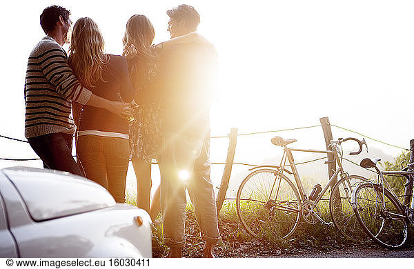 Four people standing by the side of a road  parked car and bicycles leaning against a fence  sunlight.