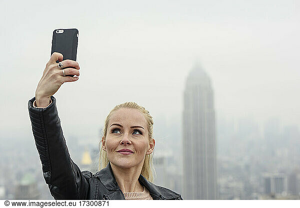Fröhliche Frau nimmt Selfie gegen moderne Wolkenkratzer in nebligen Metropole